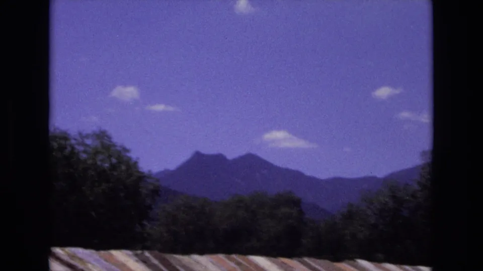 1973 Colorado 8mm frame of peaked mountain ridges under scattered clouds, with dark trees and a roofline anchoring the foreground.