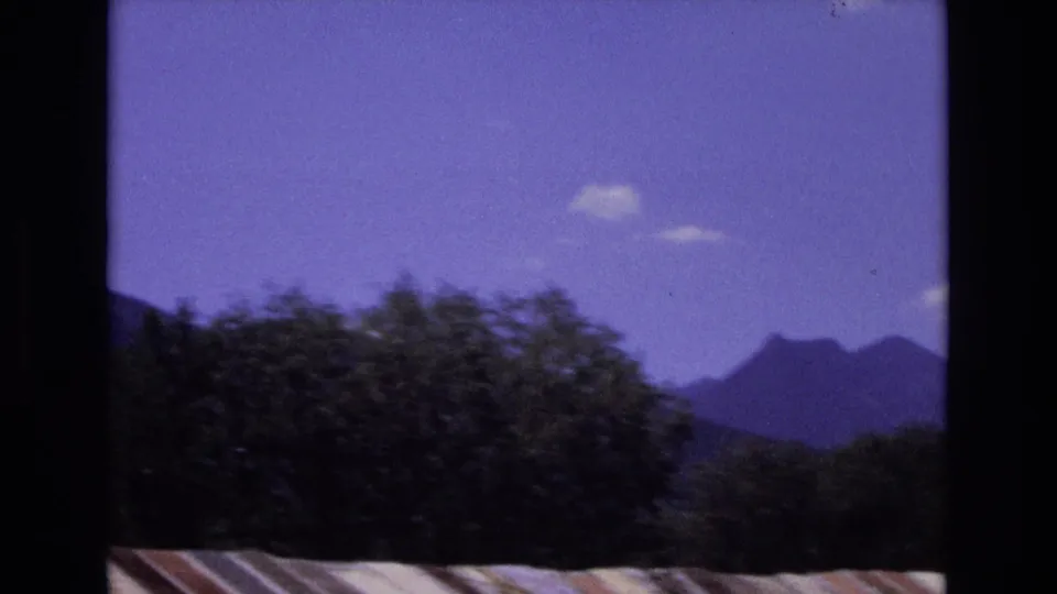 1973 Colorado 8mm frame showing distant mountain ridges and dark foreground trees beneath a saturated blue-purple sky, with a pale striped edge along the bottom.
