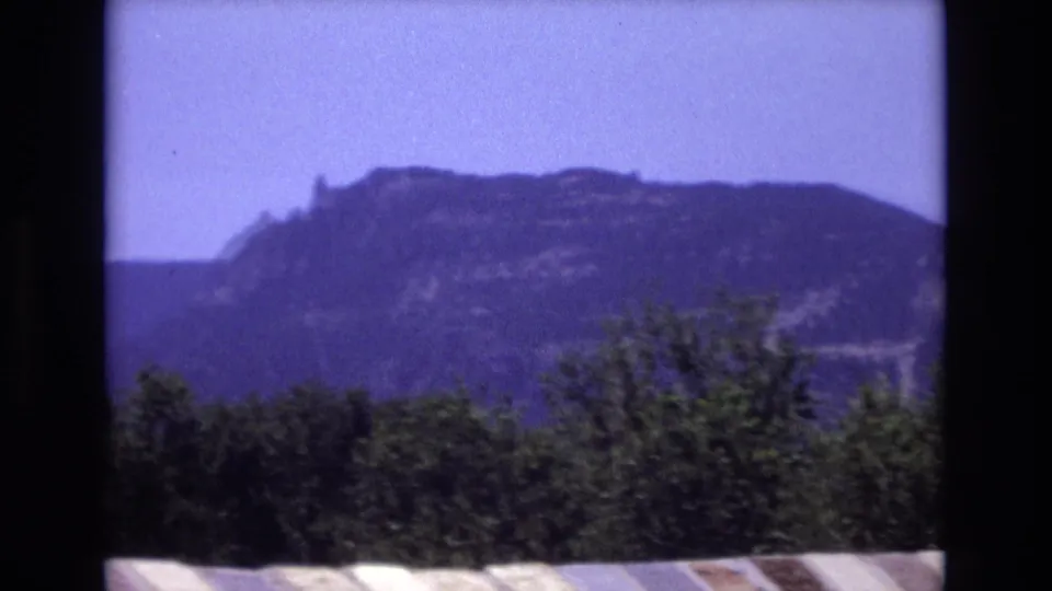 1973 Colorado 8mm frame showing a dark, rocky-looking mountain ridge beyond dense foreground trees, with saturated sky and visible film softness.