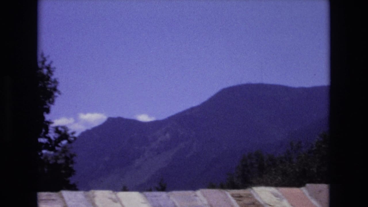 Colorado 8mm home-movie frame from 1973 showing a distant mountain ridge under a blue-purple sky, with dark trees and a pale roofline in the foreground.