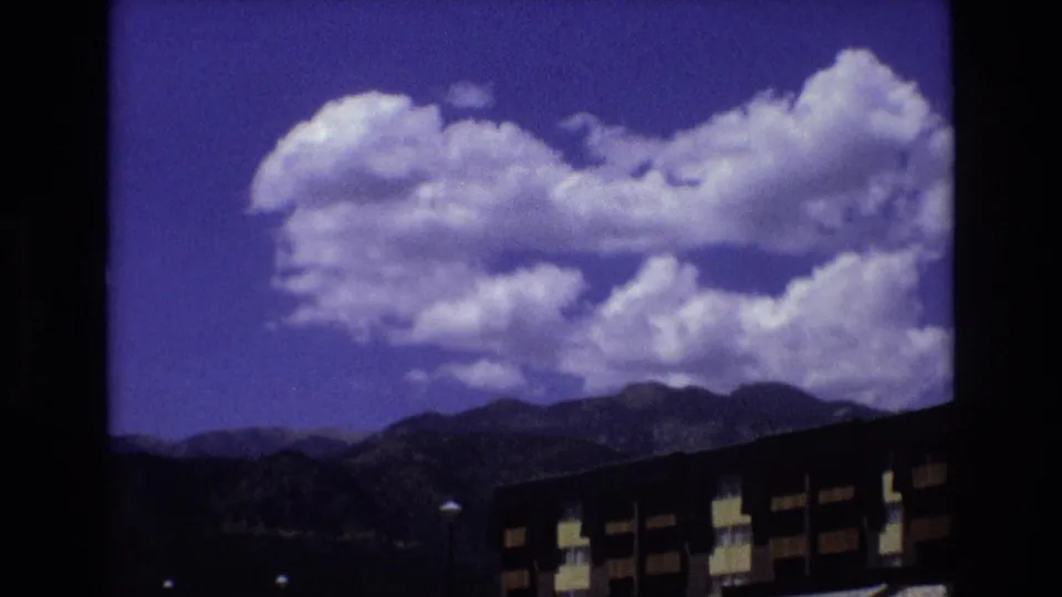 1973 Colorado home-movie view of large clouds over mountain ridges, with a dark modernist apartment building partly visible at the bottom of the frame.
