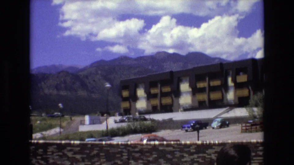 Colorado 1973 mountain-town view showing a modernist apartment complex, parking lot with period cars, cloudy ridges, and a partly visible person at the lower edge.
