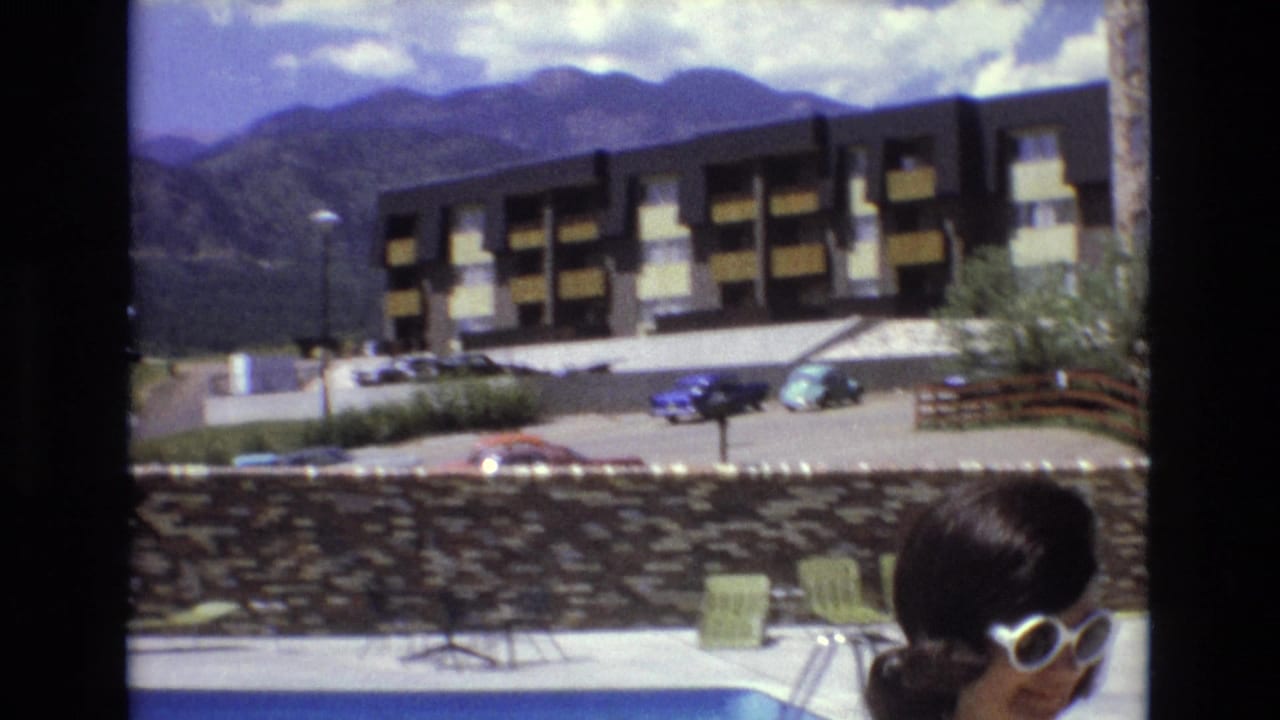 1973 Colorado home-movie frame showing a woman in white sunglasses at the edge of a swimming pool, with a hotel building, parked cars, stone wall, and mountains in the background.