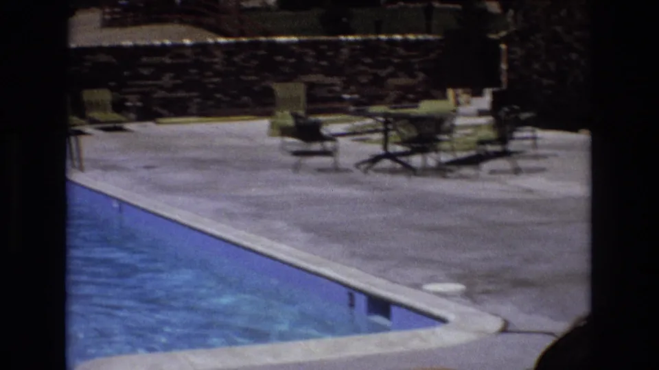 1973 Colorado resort-pool view with blue water, concrete deck, lounge chairs, patio tables, and a stone wall, with no person centered in the frame.