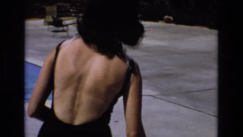 1973 Colorado home-movie frame showing a woman from behind in a black one-piece swimsuit walking away beside a resort swimming pool and concrete deck.
