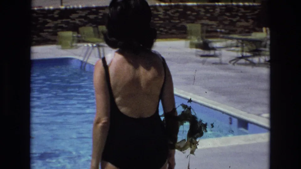 1973 Colorado home-movie frame showing a woman in a black one-piece swimsuit walking away from the swimming pool, with lounge chairs, pool steps, stone wall, and deck furniture visible.