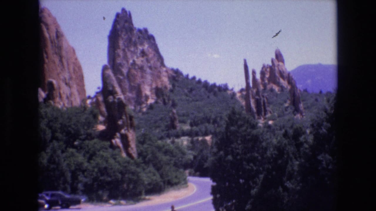 Winding road below red sandstone formations and trees with a small car at lower left.