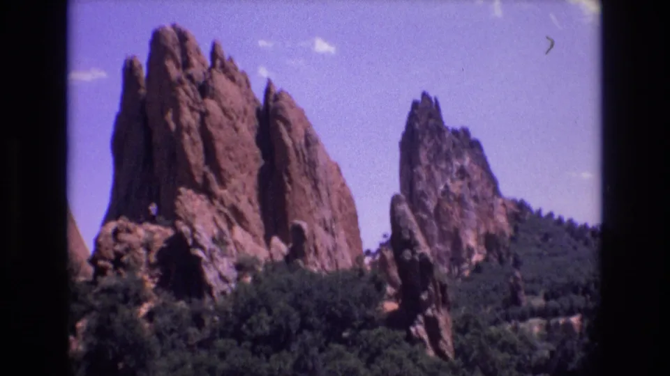 1973 Colorado home-movie frame showing tall red sandstone formations rising from green trees with additional rock ridges behind them.
