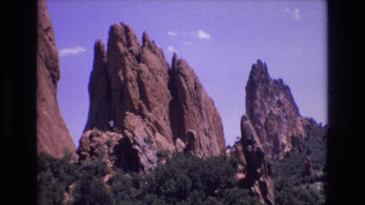 1973 Colorado 8mm frame showing red sandstone spires and green foreground vegetation under a bright blue sky, with a tiny person visible on a rock ledge.