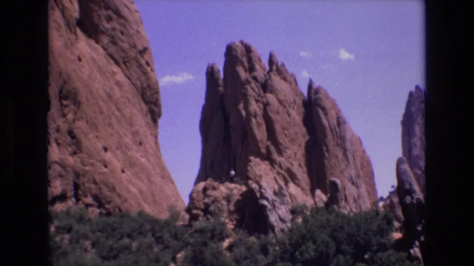 1973 Colorado 8mm frame showing a broad red sandstone wall at left, jagged spires beyond it, green shrubbery, and a distant person on a rock ledge.