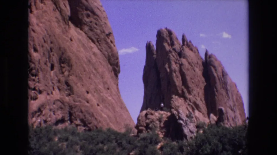 1973 Colorado frame with a close sandstone face at left, red rock spires to the right, dense green shrubs below, and a small distant person on the rocks.