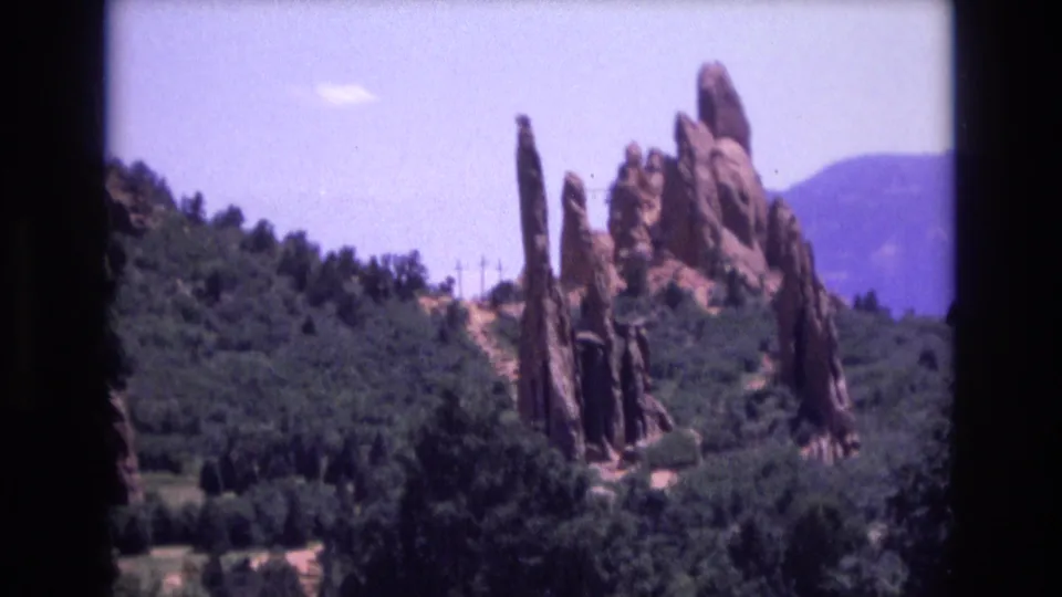 Home-movie view of Colorado sandstone formations rising from wooded slopes, with distant mountains and roadside vegetation in frame.