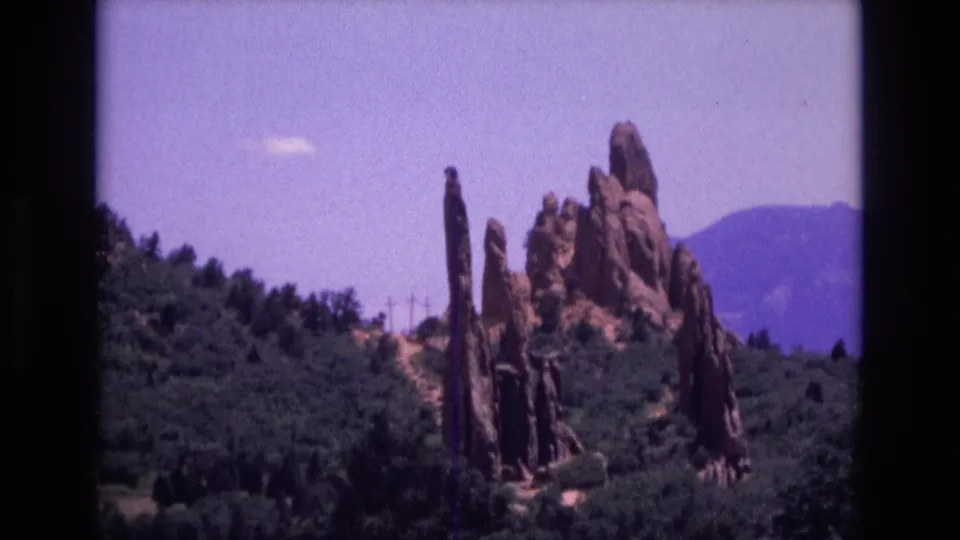 1973 Colorado frame showing tall sandstone spires and rugged rock shapes above forested terrain, with mountains faintly visible in the distance.
