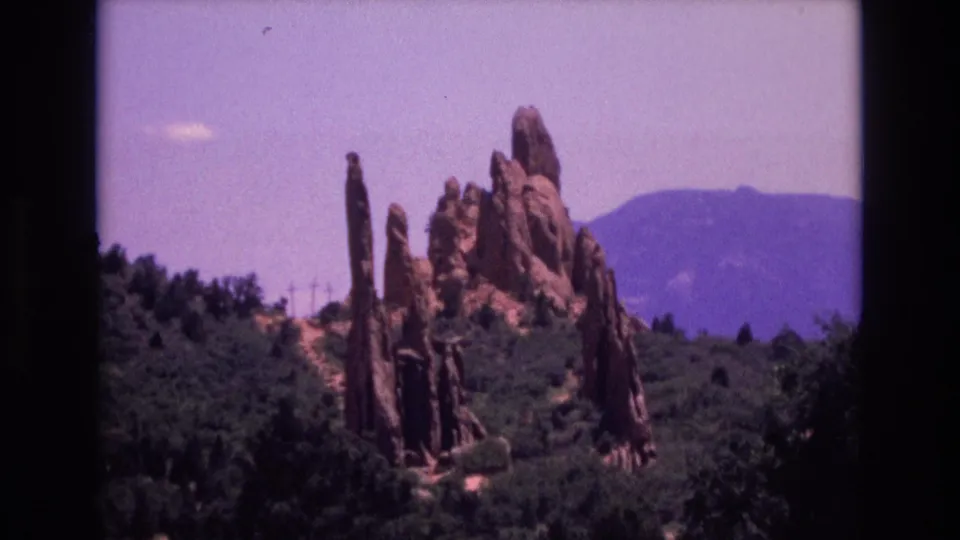 1973 home-movie frame of Garden of the Gods with red rock spires surrounded by trees and a hazy mountain backdrop.