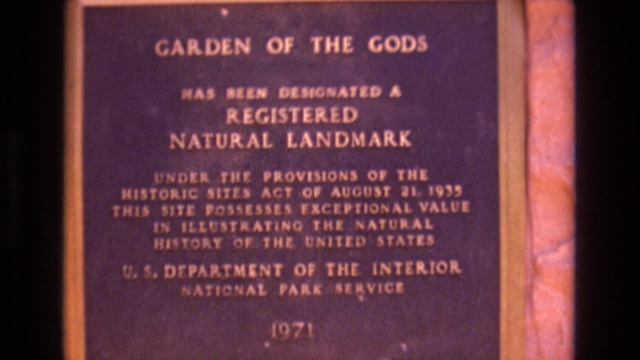 1973 Colorado 8mm frame showing a close visitor-shot view of the Garden of the Gods Registered Natural Landmark plaque with gold lettering on a dark face.