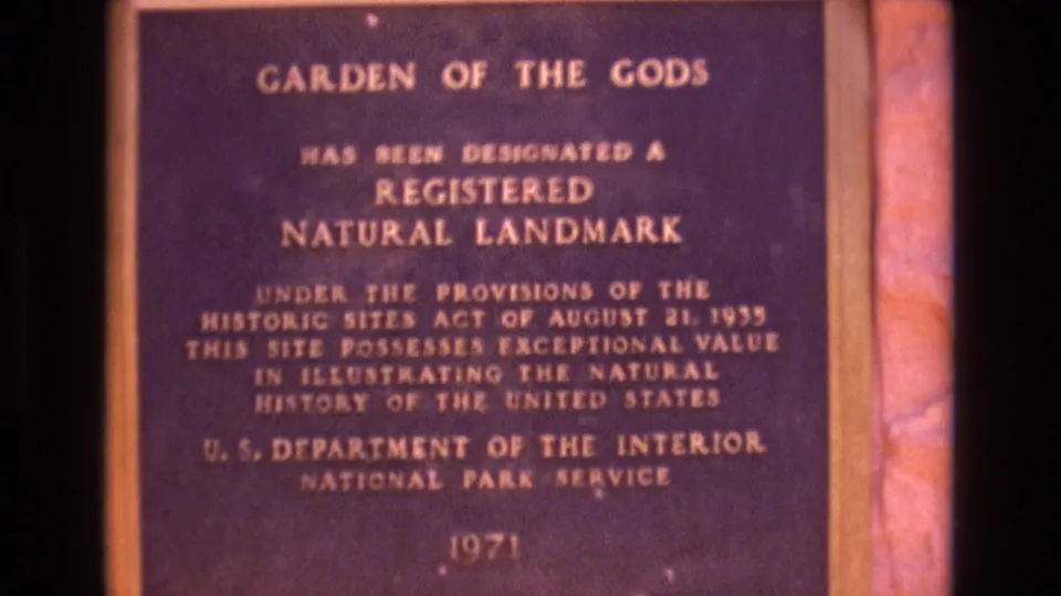 1973 Colorado 8mm frame showing a close visitor-shot view of the Garden of the Gods Registered Natural Landmark plaque with gold lettering on a dark face.