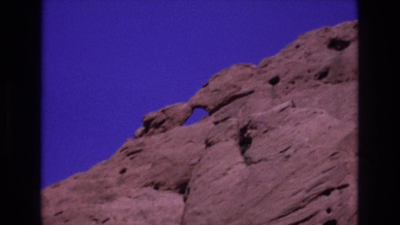 1973 Colorado 8mm frame showing reddish sandstone cliffs with an eroded opening and deep blue sky above.
