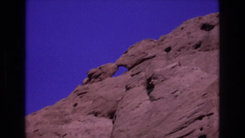 1973 Colorado 8mm frame showing reddish sandstone cliffs with an eroded opening and deep blue sky above.