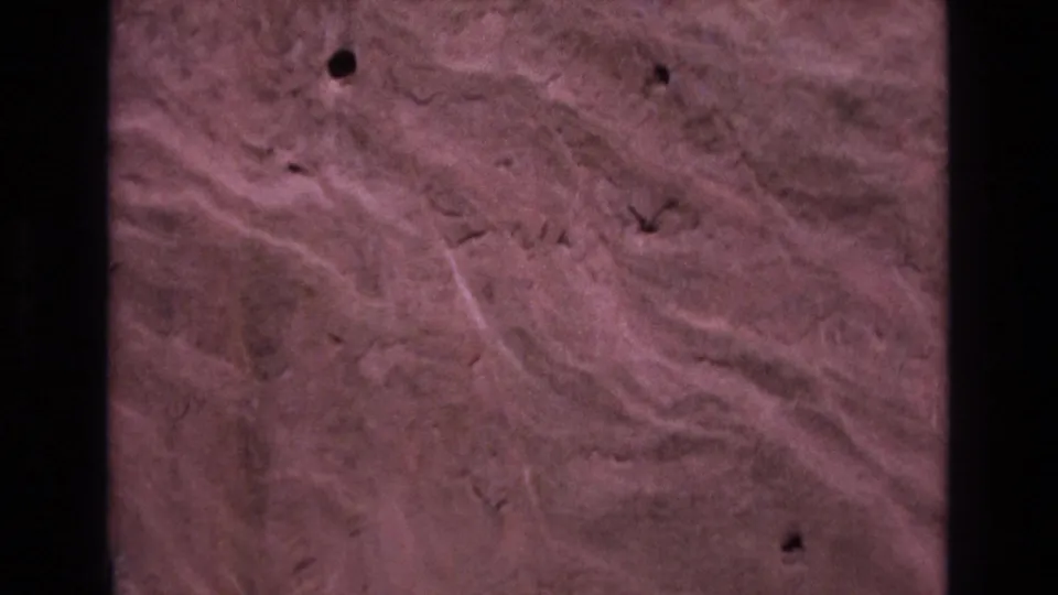1973 Colorado 8mm frame showing a close view of reddish-brown rock with one prominent dark hole, shallow cracks, and pale diagonal surface streaks.
