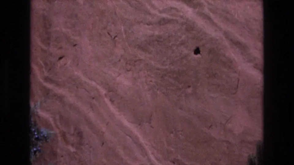 1973 Colorado 8mm frame showing a close-up of reddish rock with pale curving streaks, small pits, fine cracks, and dark film edges.