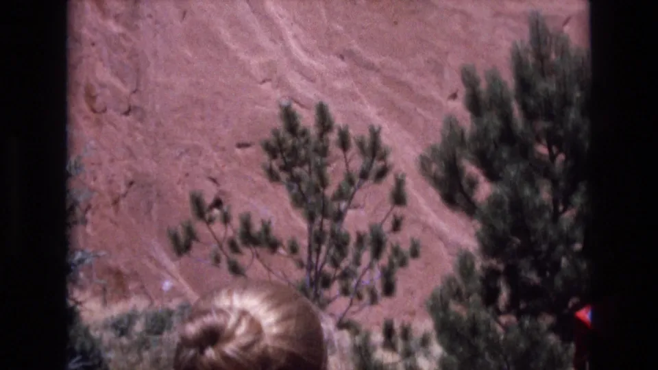 1973 Colorado 8mm frame with reddish rock, pine trees, and a partial rear view of a blonde-haired visitor entering the lower foreground.