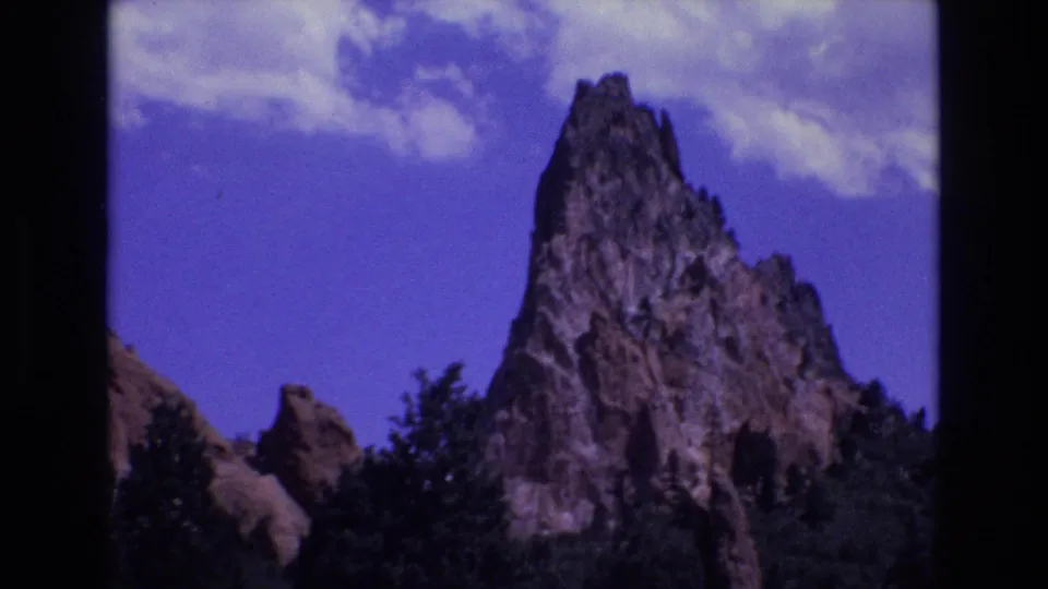 1973 Colorado 8mm frame showing a jagged red rock formation above a line of trees beneath blue sky and scattered clouds.