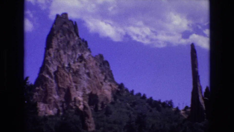 1973 Colorado 8mm frame showing a jagged rock formation with a narrow spire at right, trees in the foreground, and a bright clouded sky.