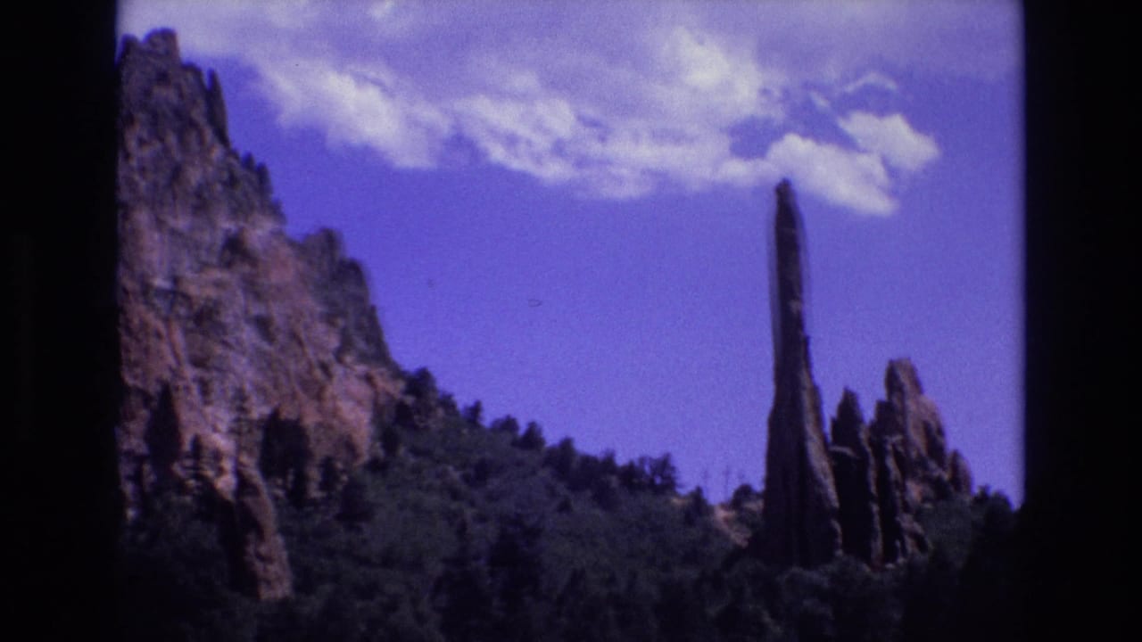 1973 Colorado 8mm frame showing a tall narrow red rock spire above dark green forest, with rugged cliff faces at left and scattered clouds overhead.