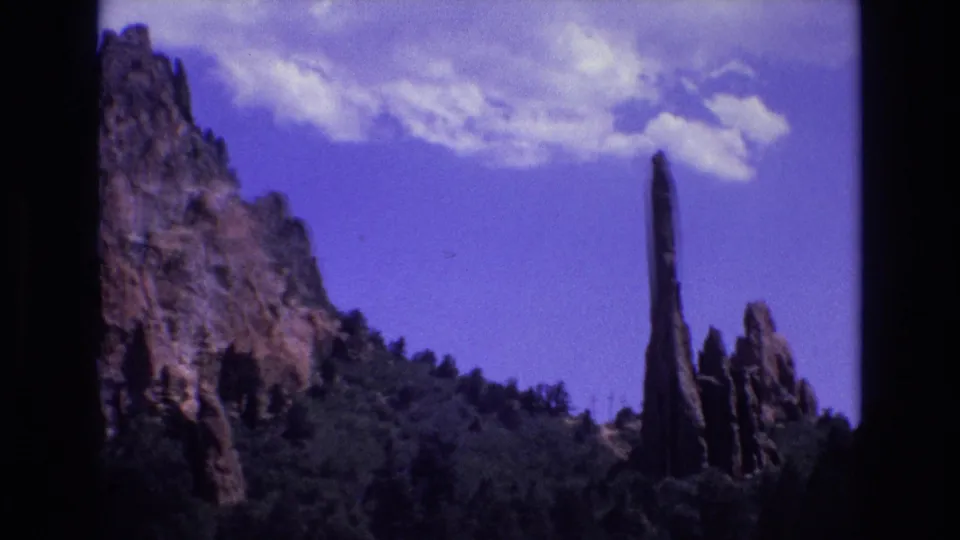 1973 Colorado 8mm frame showing a tall narrow red rock spire above dark green forest, with rugged cliff faces at left and scattered clouds overhead.