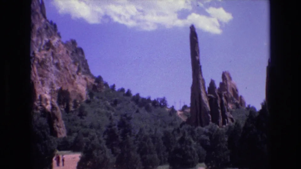 1973 Colorado 8mm frame with a tall rock spire, surrounding red cliffs, forested slopes, and very small distant visitors near a pale path at the bottom.