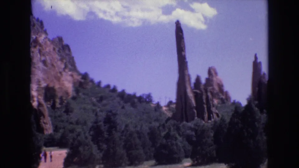 1973 Colorado 8mm frame showing red rock formations above dense green trees, with a pale path and tiny visitors near the lower left for scale.