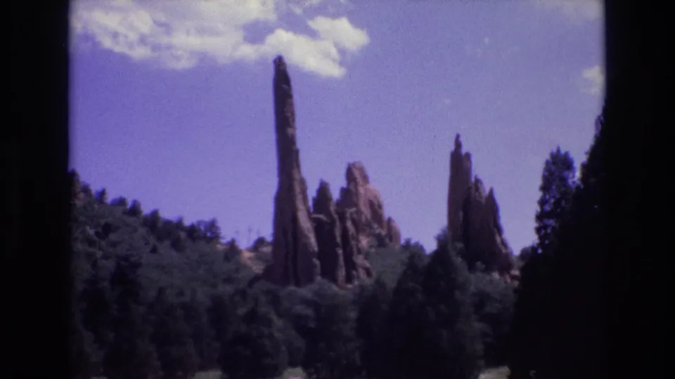 1973 Colorado 8mm frame showing several narrow red rock spires rising from forested terrain beneath a blue-purple sky, with no identifiable faces or signage visible.