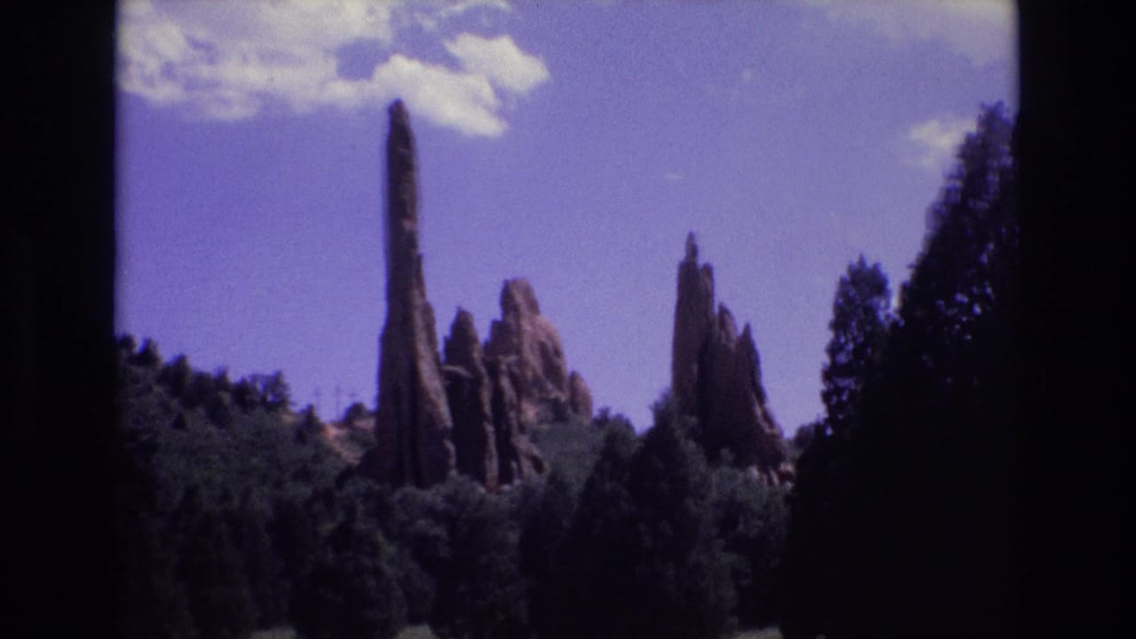 Tall red rock spires rise behind a dark conifer forest under a blue sky with clouds.