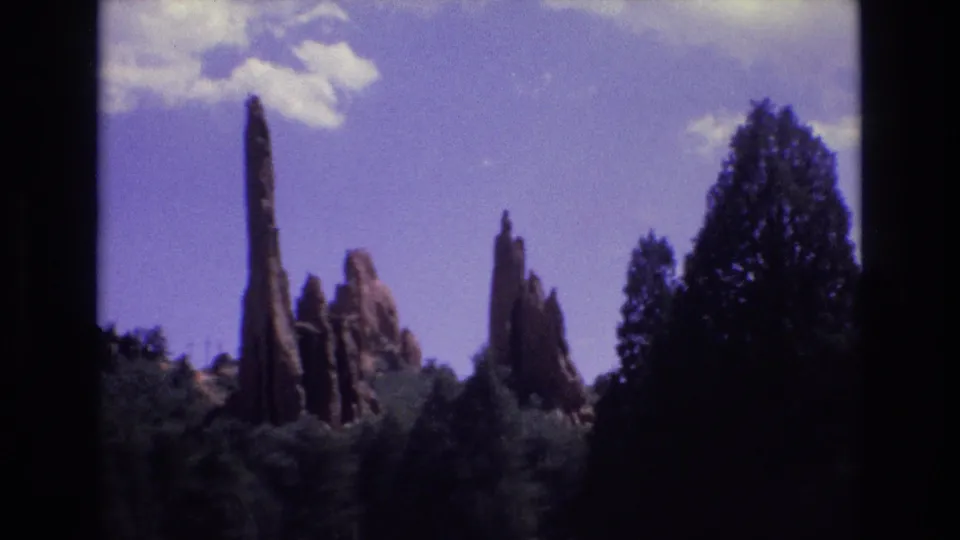 1973 Colorado 8mm view of vertical red rock formations rising beyond conifers, framed by a dark tree silhouette and soft blue sky.