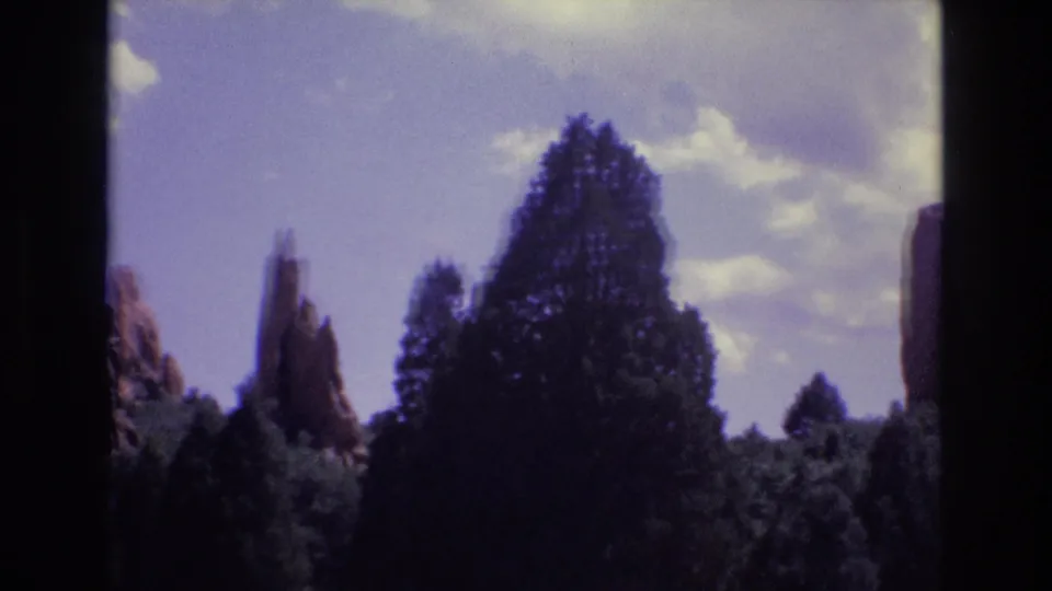 1973 Colorado 8mm frame with a central dark conifer obscuring part of the red rock spires, beneath pale clouds and a soft blue sky.