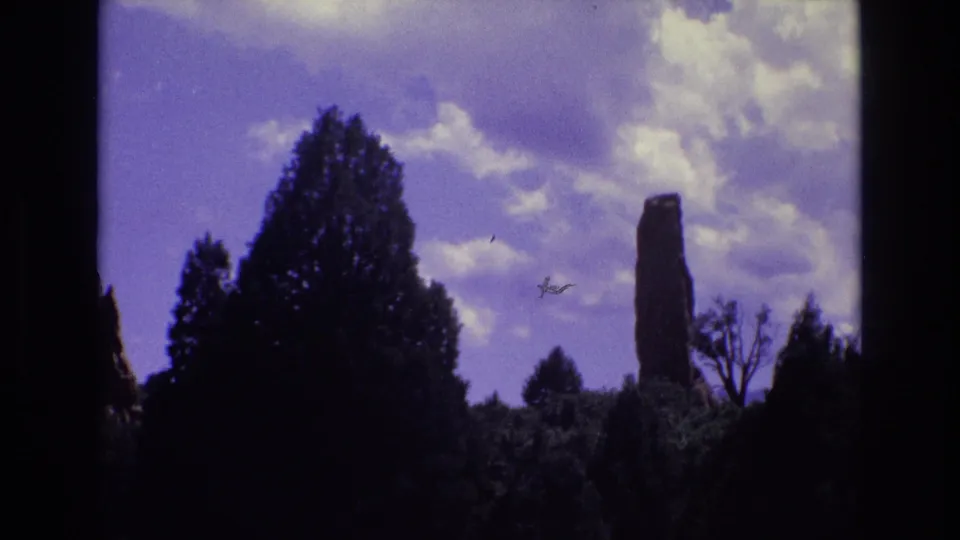 1973 Colorado 8mm frame showing dark conifers, a tall reddish rock column at right, scattered clouds, and a small speck or scratch in the sky area.