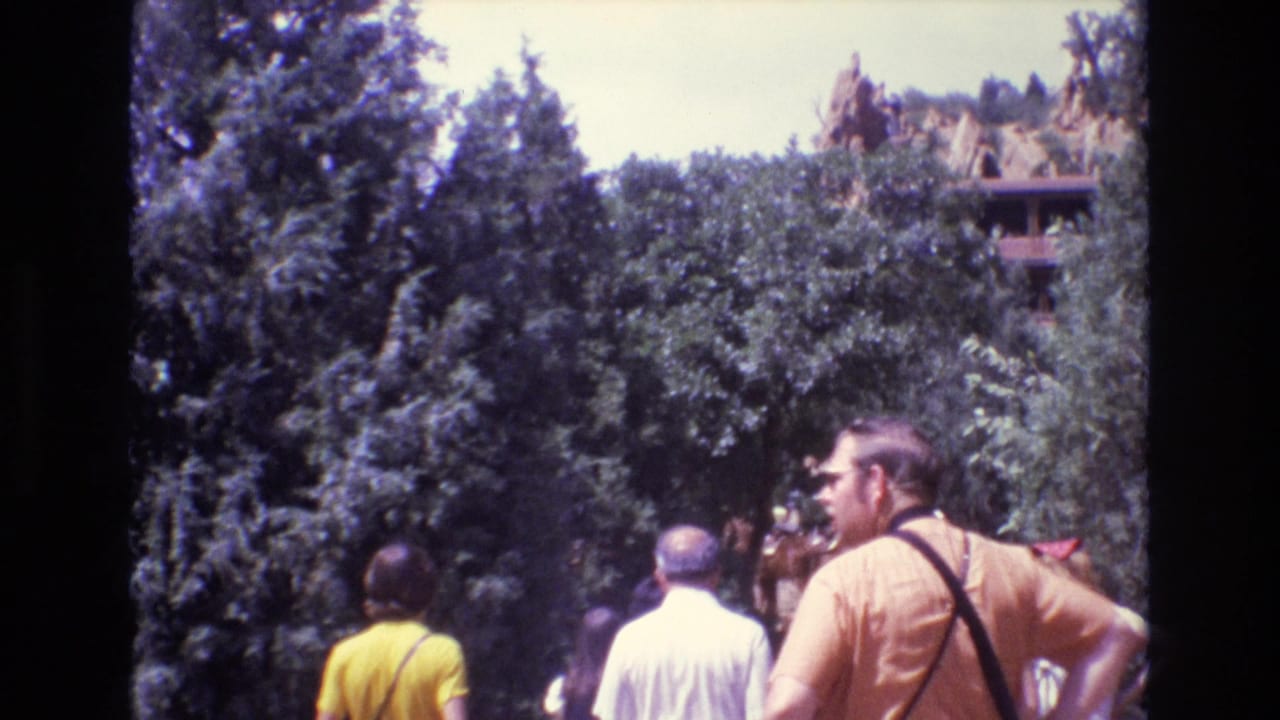 Tourists walk through trees toward tipi-like structures on a hillside.