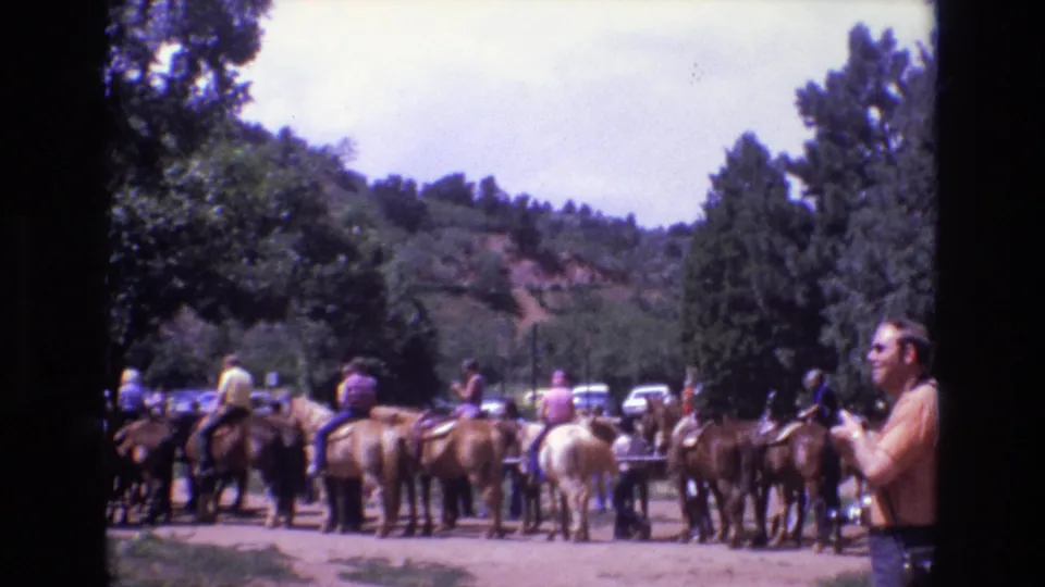 1973 Colorado 8mm frame showing riders on horses in a wooded recreation area as an adult man at right holds a camera toward the group.