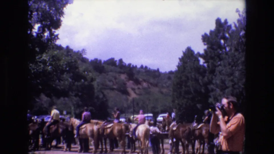 1973 Colorado home-movie frame with a group of tourists on horseback viewed from behind, trees and hillside beyond, and a camera-holding adult in the right foreground.