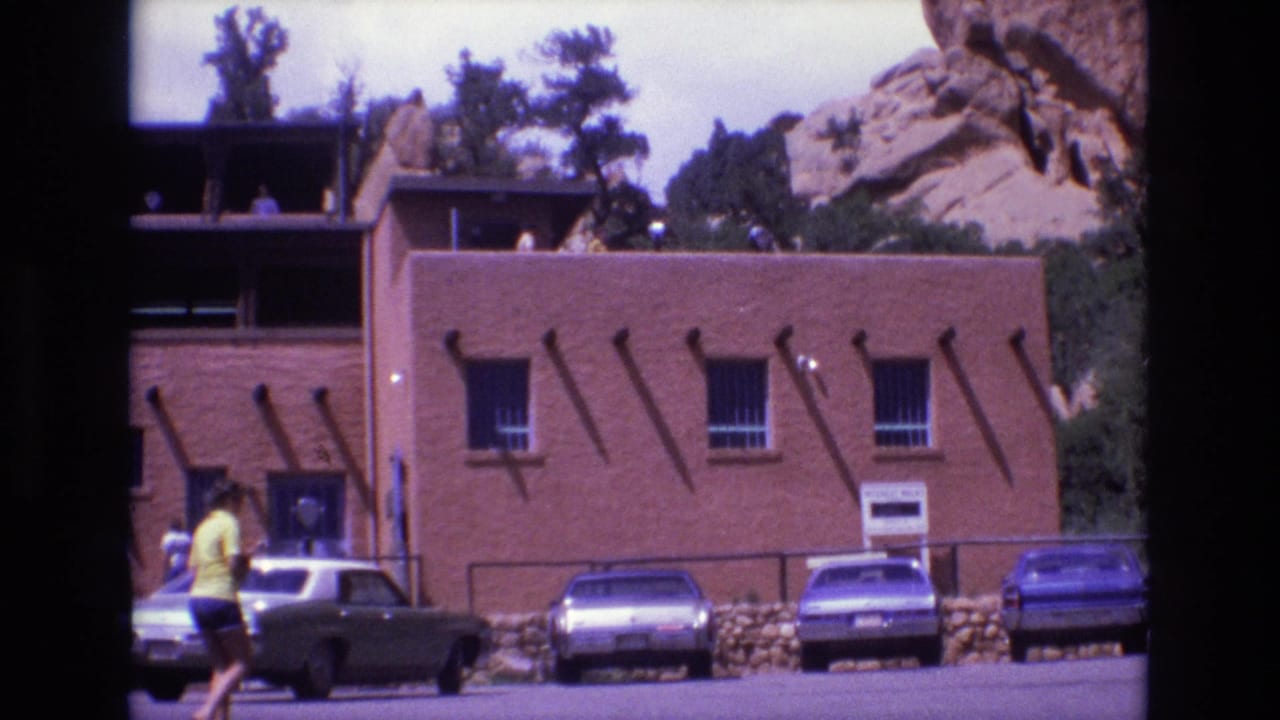Adobe-like building with parked cars, pedestrians, and red rock terrain behind it.