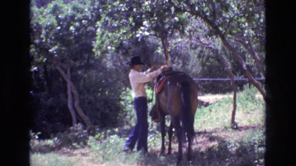 1973 Colorado 8mm frame showing a man in a black hat adjusting or freeing reins near a brown horse's head in a shaded green clearing.