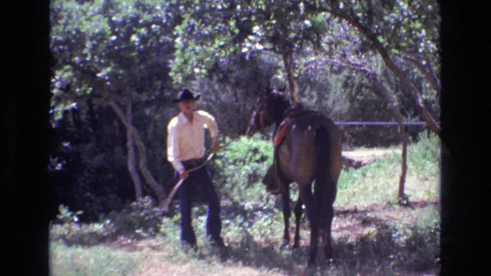 1973 Colorado 8mm frame showing a man in a black hat stepping away with reins or lead line as the brown horse stands among trees and brush.