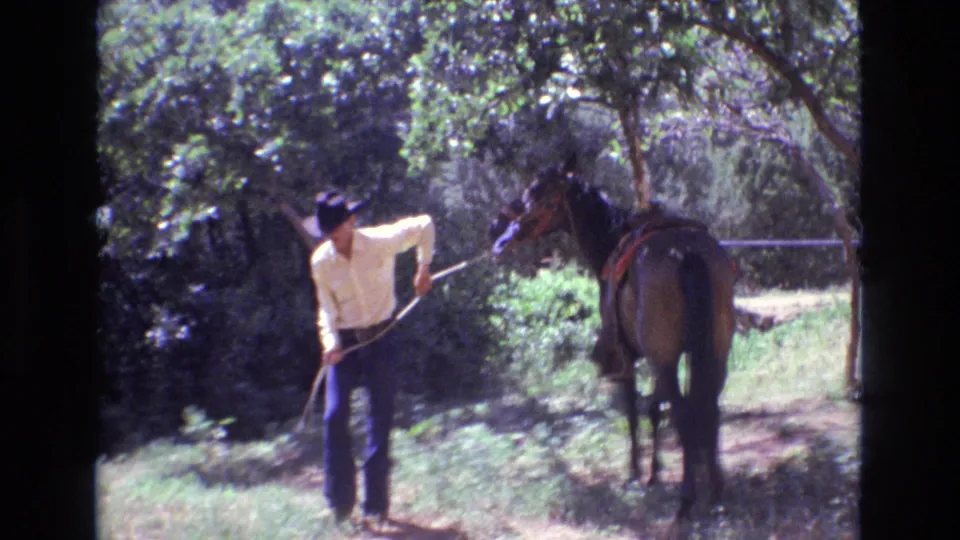 In 1973 Colorado 8mm footage, an adult man in a dark cowboy hat and light shirt leads a brown saddled horse through a sunlit wooded area.
