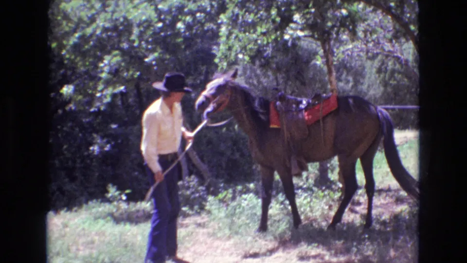 An adult man in a cowboy hat holds the reins beside a brown horse with a red saddle blanket in a wooded Colorado setting from 1973 home-movie footage.