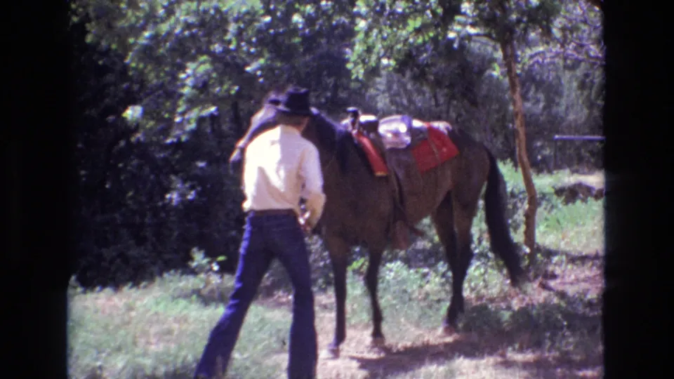 Seen from behind, an adult man in a dark hat and light shirt approaches or handles a saddled horse in a sun-dappled wooded area in Colorado, 1973.