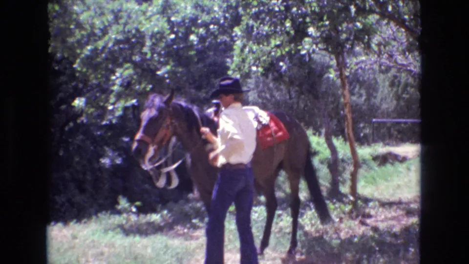 An adult man in a cowboy hat holds the reins as the saddled brown horse turns near him in bright, tree-filtered Colorado light in 1973 8mm footage.
