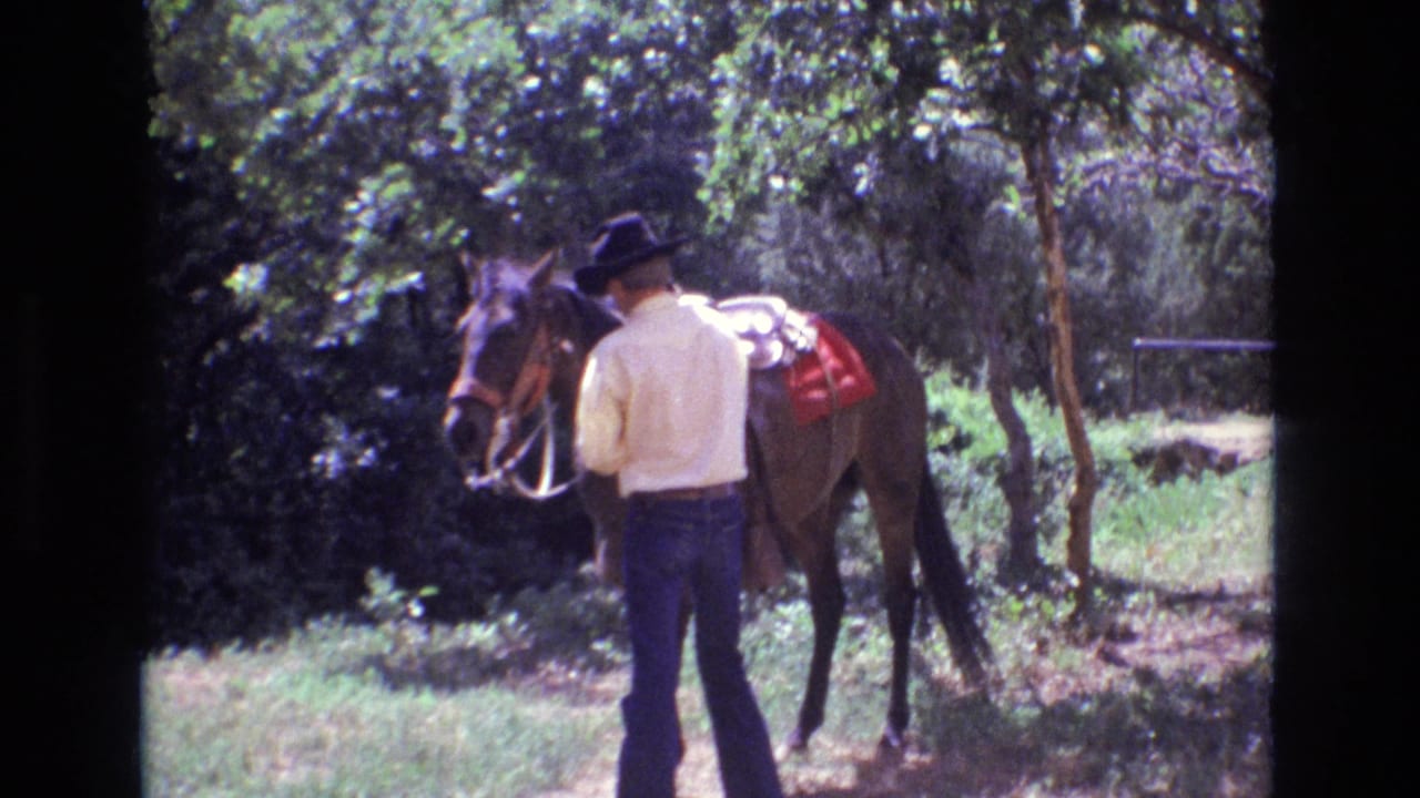 In 1973 Colorado home-movie footage, an adult man in a black cowboy hat stands beside a saddled brown horse in a wooded area.