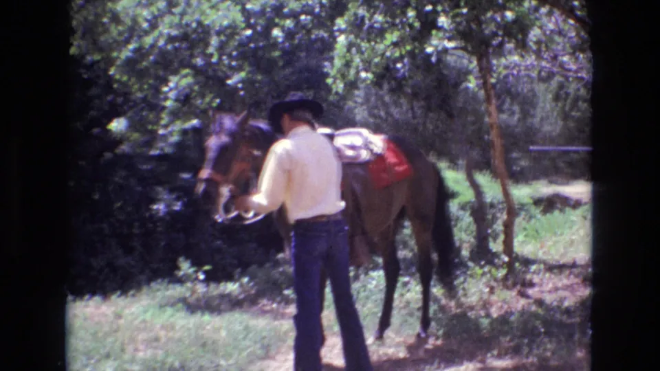 An adult cowboy appears to handle the reins or tack of a brown horse along a dirt path in wooded Colorado surroundings.