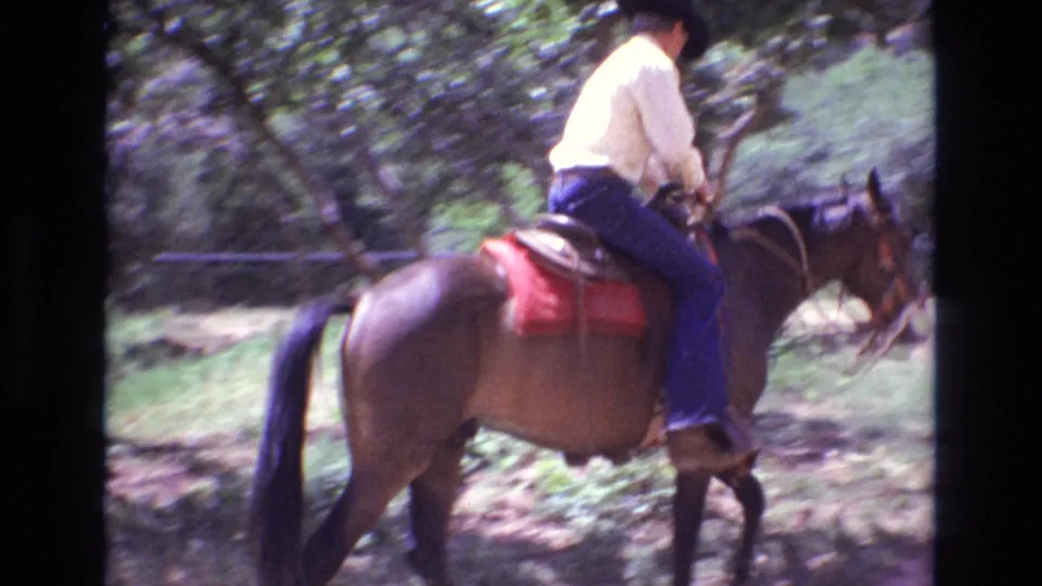 An adult cowboy rides a brown horse through a wooded Colorado setting, with the red saddle blanket visible as the horse moves along the path.