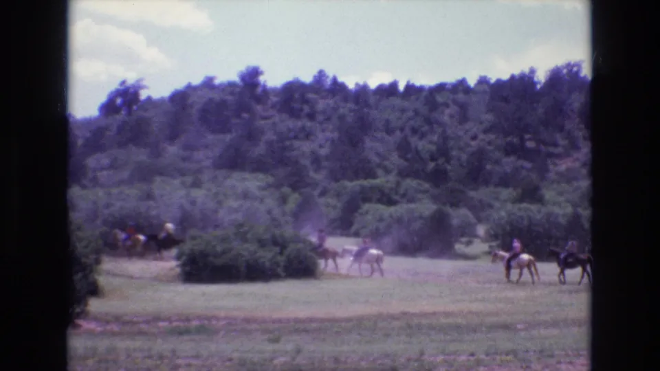 A line of horseback riders continues across the field in Colorado, with dust near the path and dark 8mm frame dividers around the scene.